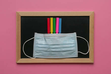 Face mask on top of colored pens and blackboard. Pink background.