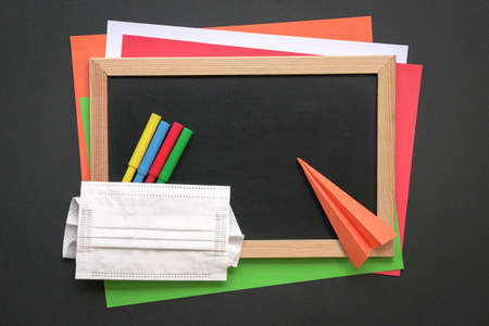 Face mask on top of black board and colored papers with magic coloring pens and paper plane. Top...