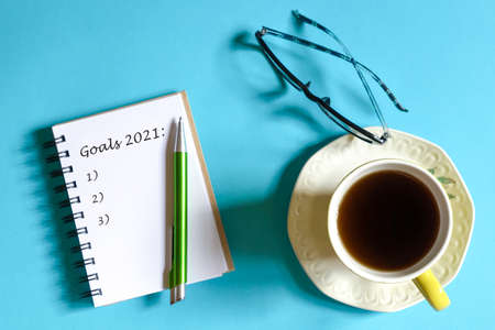 Notepad with words, Goals 2021 and pen, Reading glasses and cup of coffee on blue background. Top...