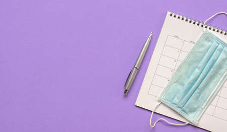 Calendar with medical face mask and pen on purple background. Top view. Copy space.