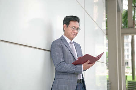 Asian businessman leaning against whilte wall and looking at his diary.
