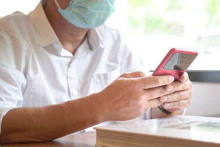 Man with face mask sitting next to windows texting on his phone. Closeup view.の写真素材