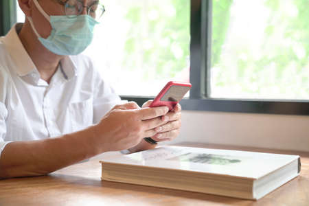 Man with face mask sitting next to windows with a book in front of him, and texting on his phone.の写真素材