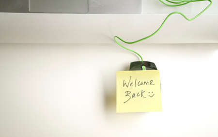 Welcome back with smiley written on yellow paper stick on computer mouse on top of office table.の写真素材