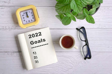 New year goals 2022 on desk. Top view with coffee, notepad, clock, eyeglasses and green plant.