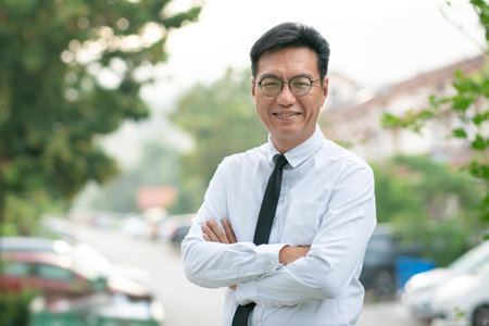 Well dressed mature Asian businessman with arms crossed, smiling confidently. Urban residential property at the background.の写真素材