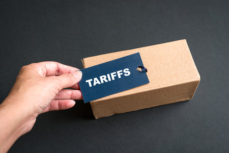Hand holding tag with tariffs message, on top of cardboard package.の写真素材