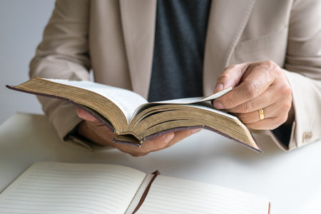 Preacher holding an open old rustic bible in his hands, with a notepad on top of desk.