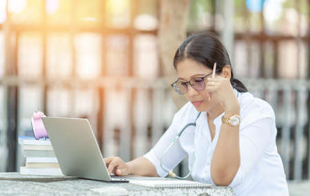 Portrait of female student sitting at the park and using laptop ,  writing thesis with serious face expression. Young asian woman working on her home assignmentの写真素材