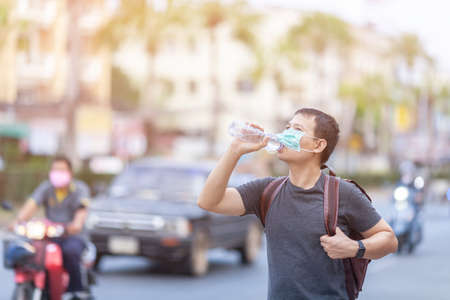 A young asian man standing drinks water with bottle of water in his hands, waiting for the car at road, travel, and vacation ideasの写真素材