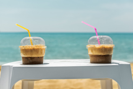 Two ice frappe coffee on white plastic coffee table in plastic transparent cups, with yellow and magenta straws, on summer day at beach with blue sea and yellow sand on background.の写真素材