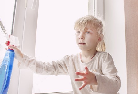 A little blonde in white top and blue leggings standing in the room sprinkles on the window, a remedy.の写真素材