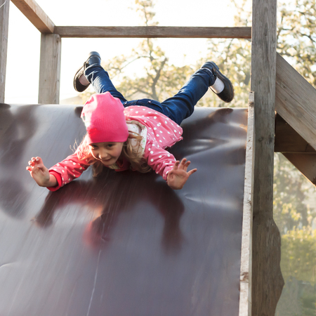 Girl in a red jacket. In blue pants and in dark shoes slides down from a wooden hill in retro styleの写真素材