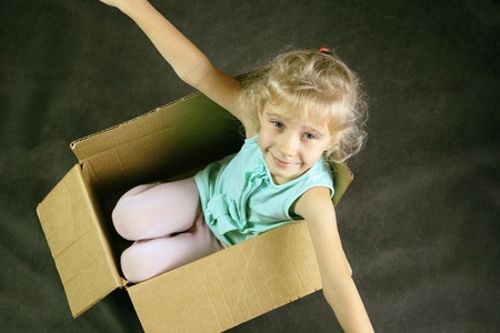 The little blonde smiles inside a cardboard box. shows us hands. dark background. toning.
