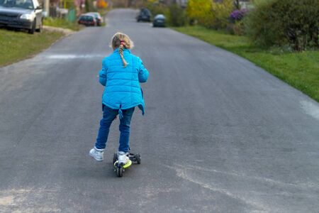 back view. girl in a blue jacket riding a scooter. blond hair color. she rolls her foot on the board