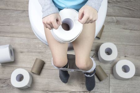 the child sits on a white toilet. in his hands is toilet paper. there are a lot of rolls on the floor. close-upの写真素材