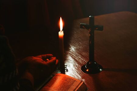 Wooden table in retro style. On it is a candle and a cross. Hands of an old woman over a book. Palms folded.の写真素材