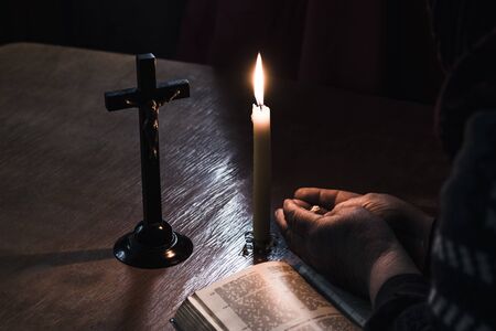 Wooden table in retro style. On it is a candle and a cross. Hands of an old woman over a book. Palms...