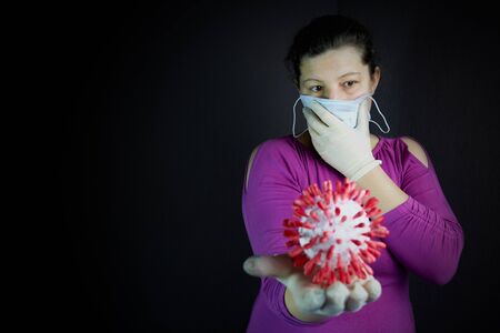 A dark room. In it, a girl with dark hair with one hand holds a medical mask at her mouth. On the other hand is a model of the coronavir molecule.の写真素材