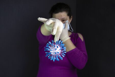 A dark room. In it, a girl with dark hair with one hand holds a medical mask at her mouth. On the other hand is a model of the coronavir moleculeの写真素材