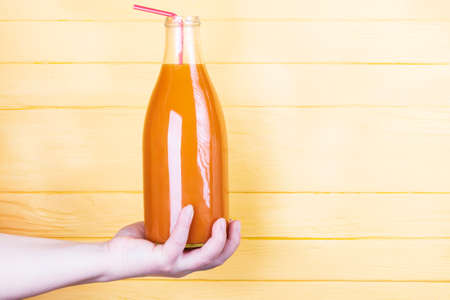 yellow, wooden background. a glass bottle with an orange liquid inside. the juice of tangerine and carrots, as well as pumpkins, is held by a female hand.の写真素材