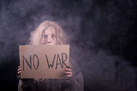 lots of smoke. A frustrated little girl, a homeless child with a dirty face, raises a banner with the inscription No war on a black background. Crisis, peace, stop aggression, child against warの写真素材