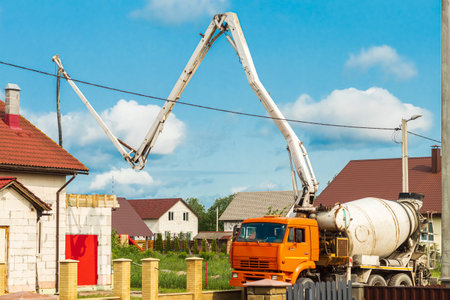 A concrete truck with a long boom is pouring fresh cement into a foundation at a construction site surrounded by houses under a clear blue sky....の写真素材