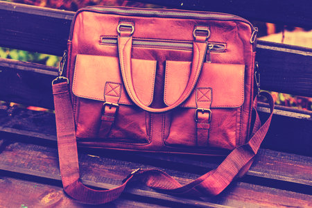 Stylish brown leather bag resting on a wooden bench in a quiet outdoor setting during the dayの写真素材