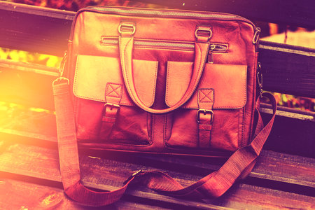 Stylish brown leather bag resting on a wooden bench in a quiet outdoor setting during the dayの写真素材