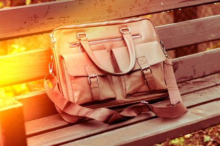 Stylish brown leather bag resting on a wooden bench in a quiet outdoor setting during the dayの写真素材