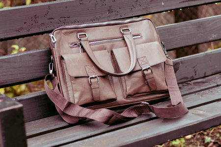 Stylish brown leather bag resting on a wooden bench in a quiet outdoor setting during the dayの写真素材