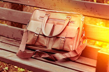 Stylish brown leather bag resting on a wooden bench in a quiet outdoor setting during the dayの写真素材