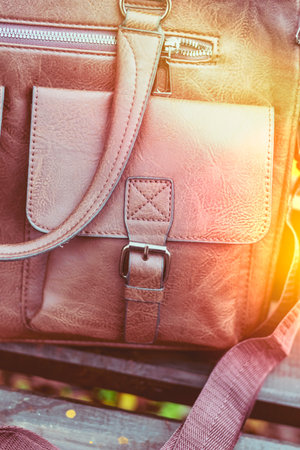 Stylish brown leather bag resting on a wooden bench in a garden setting during autumn with colorful leaves scattered aroundの写真素材
