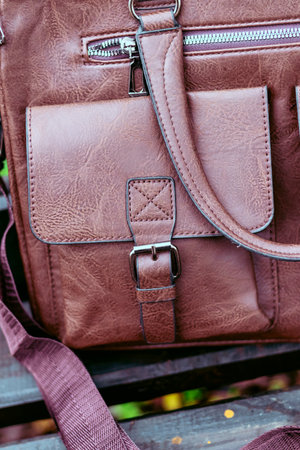 Stylish brown leather bag resting on a wooden bench in a garden setting during autumn with colorful leaves scattered aroundの写真素材