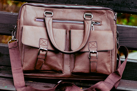Stylish brown leather bag resting on a wooden bench in a quiet outdoor setting during the dayの写真素材