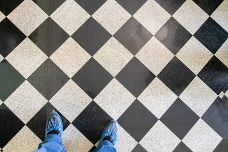 A person stands on a checkered black and white floor. The blue shoes are visible, while the surrounding area is well lit, suggesting it is daytime.