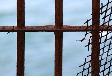 Rusty metalic fence with holes.   Fence bars  and  rests of rusty net, the sea behind.の写真素材