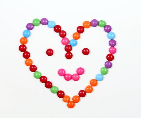 Smiling heart made from colored smarties. Chocolate candies in a shape of heart with smile on face, white background,の写真素材