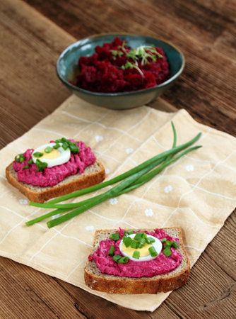 Beetroot mixed with goat cream cheese spreaded on whole grain bread. Decorated with slice of egg and green onion. Red beetroot salad and herbs at back.の写真素材