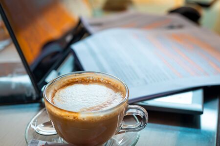 A cup of cappuccino with paperworks resting on a laptop in the backdrop. Working on a project or assignment concept.の写真素材
