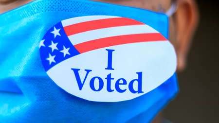 Voting during Coronavirus Pandemic concept. Man puts I Voted sticker with American Flag on the face mask.の写真素材