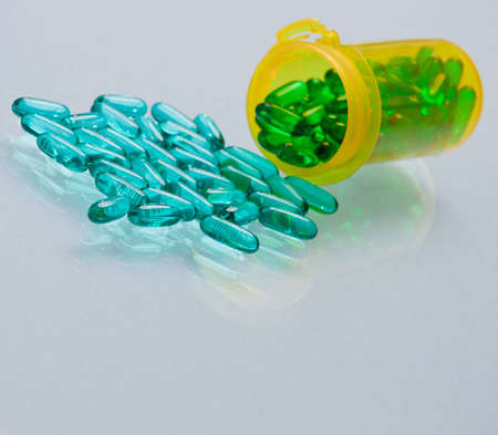 A close up of a bunch of turquoise pills wtih a yellow vial and a white glass background.
