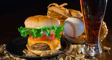 A close up of a big hamburger surrounded with peanuts, beer and a baseball glove and ball.
