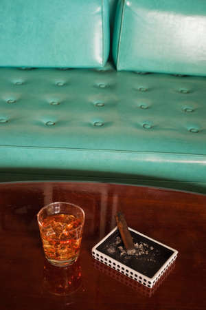 A shot of a old wooden coffee table with a dice pattern ash tray and cigar with a low ball glass of whiskey and a turquoise couch in the background.の写真素材