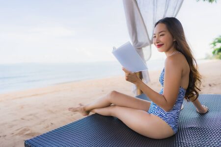 Portrait beautiful young asian woman reading book with happy smile relax in lounge bed chair on the beach sea ocean for leisure travel in vacationの写真素材