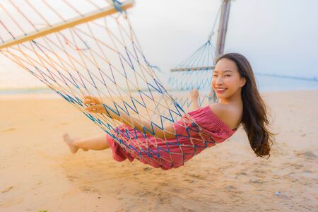 Portrait beautiful young asian woman sitting on the hammock with smile happy neary beach sea and ocean for leisure travel and vacation at sunset timeの写真素材