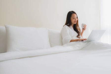 Portrait beautiful young asian women with coffee cup and computer laptop on bed in bedroom interiorの写真素材