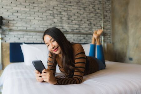 Young asian woman using mobile smart phone on bed in bedroom interiorの写真素材
