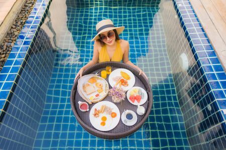 Portrait beautiful young asian woman happy smile relax with breakfast floating around swimming pool in hotel resortの写真素材