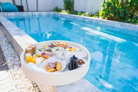 Floating breakfast set in tray with fried egg omelette sausage ham bread fruit milk juice coffee and other around swimming poolの写真素材